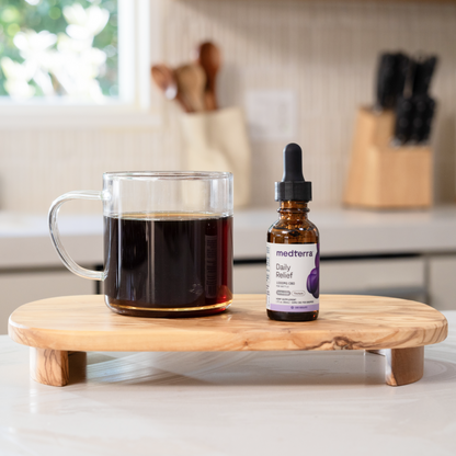 A glass mug filled with a dark beverage and a small bottle labeled "Medterra Daily Relief CBD Isolate Oil" are placed on a wooden stand in a kitchen setting. The countertop in the background features utensils and a knife block.