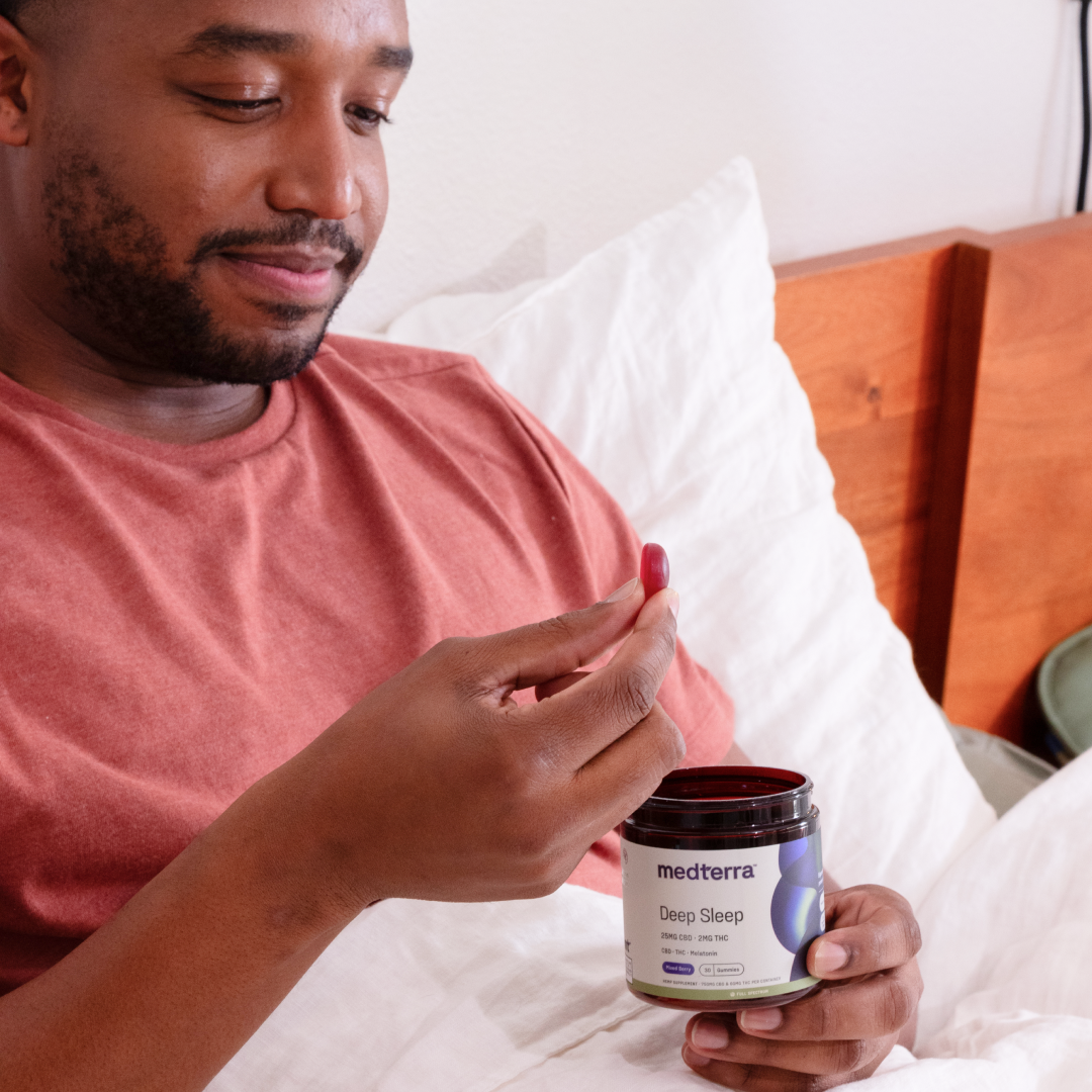 A person wearing a red shirt is sitting up in bed, holding a small red gummy in one hand and a jar labeled "Medterra Deep Sleep CBD Gummies" in the other. The jar boasts Full Spectrum Hemp Extract and melatonin, suggesting the person is preparing for a restful night’s sleep.