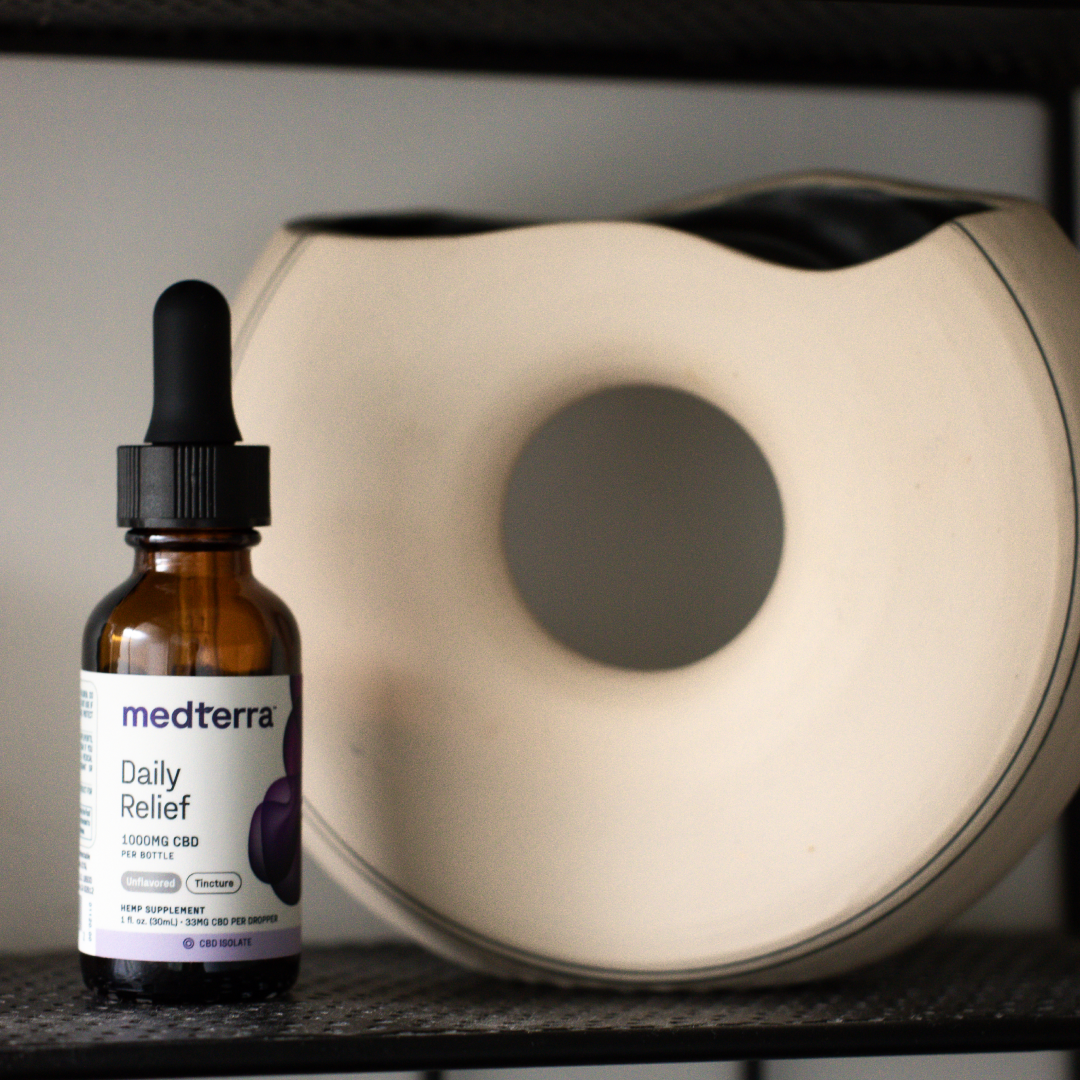 A bottle of Medterra Daily Relief CBD Isolate Oil, 1000mg, is placed on a shelf beside a minimalist ceramic vase with a circular opening. The bottle features a dropper lid and a label detailing the product information. The scene is well-lit, with the background tastefully blurred.